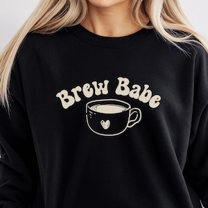 Peut inclure: Sweat-shirt noir avec l'inscription "Brew Babe" dans une police rétro au-dessus d'un dessin de tasse à café avec un cœur. Le sweat-shirt est de style col rond et a des manches longues.