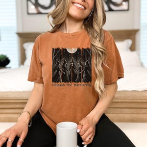 Peut inclure: T-shirt orange br&ucirc;l&eacute; avec un motif de trois sorci&egrave;res dans une for&ecirc;t sous la pleine lune. Le texte "Unleash The Matriarchy" est imprim&eacute; en dessous. Le t-shirt a des manches courtes et une coupe d&eacute;contract&eacute;e.