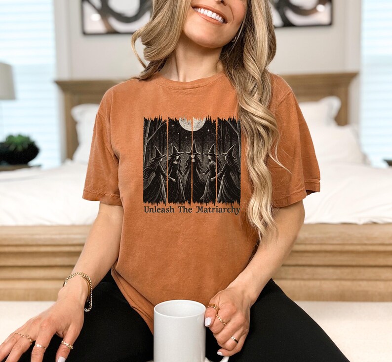 Peut inclure: T-shirt orange br&ucirc;l&eacute; avec un motif de trois sorci&egrave;res dans une for&ecirc;t sous la pleine lune. Le texte "Unleash The Matriarchy" est imprim&eacute; en dessous. Le t-shirt a des manches courtes et une coupe d&eacute;contract&eacute;e.