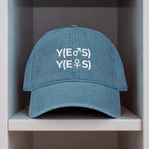 May include: A blue baseball cap with the text "Y(E♂S) Y(E♀S)" in white. The cap has a curved brim and a button on top. The hat is made of a soft material and is a casual accessory.