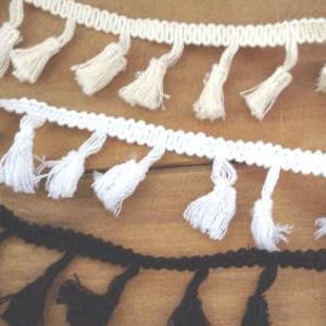 May include: Three strips of decorative fringe trim featuring small hanging tassels. The top strip is cream, the middle strip is bright white, and the bottom strip is black. Each trim consists of a braided header with evenly spaced yarn tassels.
