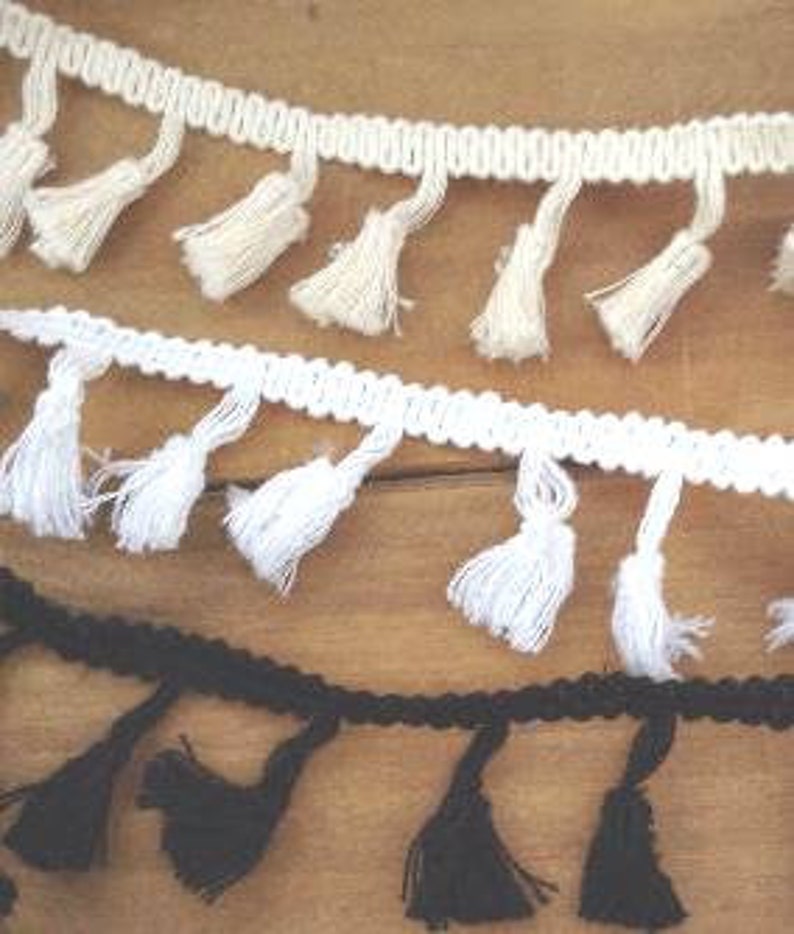 May include: Three strips of decorative fringe trim featuring small hanging tassels. The top strip is cream, the middle strip is bright white, and the bottom strip is black. Each trim consists of a braided header with evenly spaced yarn tassels.