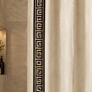 May include: A beige shower curtain featuring a vertical black and gold Greek key pattern border along the side. The curtain is suspended from a dark metal rod with matching rings, displaying a textured fabric finish throughout the panel.