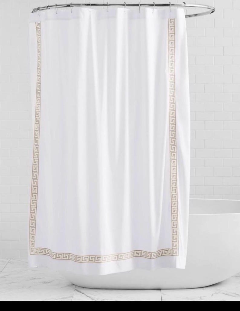 May include: A white shower curtain featuring a vertical and horizontal border with a tan-colored Greek key pattern. The curtain hangs from a circular metal rod and is made of a smooth fabric that drapes in soft, vertical folds.