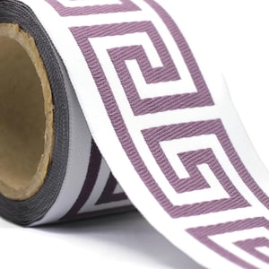 May include: A roll of woven fabric ribbon featuring a repeating white Greek key pattern set against a solid mauve background. The ribbon has a textured surface and is wound around a cylindrical cardboard core, showing a consistent design.