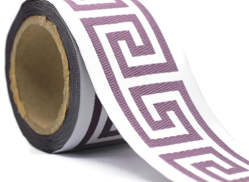 May include: A roll of woven fabric ribbon featuring a repeating white Greek key pattern set against a solid mauve background. The ribbon has a textured surface and is wound around a cylindrical cardboard core, showing a consistent design.