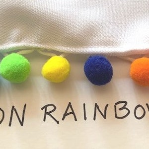 May include: A white fabric edge trimmed with a row of small, round, multi-colored pom-poms in shades of pink, blue, green, yellow, dark blue, orange, and magenta. The text NEON RAINBOW is printed in black capital letters below the pom-pom trim.
