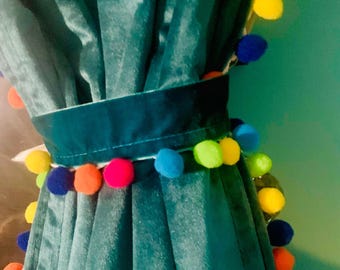Può includere: Tenda in velluto verde acqua con un fermatenda abbinato adornato con un bordo di pompon colorati. La tenda è drappeggiata, mostrando morbide pieghe e consistenza. I pompon sono nelle tonalità di blu, giallo, arancione e rosa.