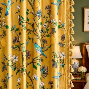 May include: Mustard yellow curtain with a botanical print featuring hummingbirds, flowers, and pears. The curtain has a gold tassel trim and is hanging on a dark wooden rod. The design includes shades of blue, green, and white.