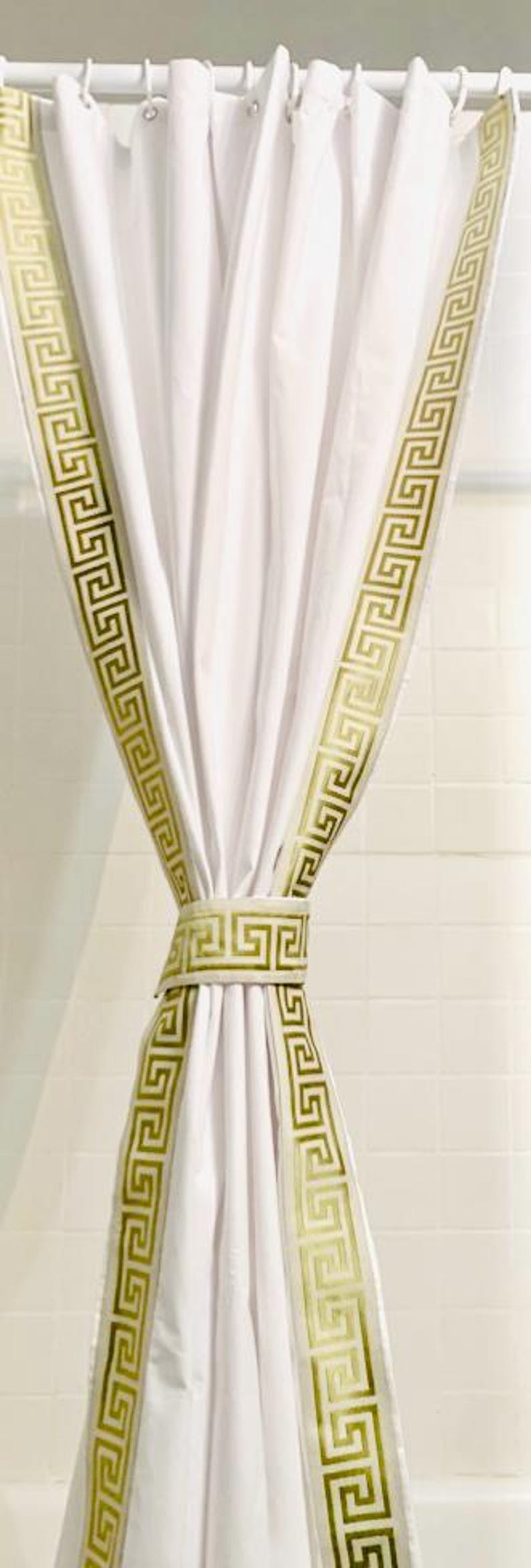 May include: A white shower curtain featuring a vertical gold Greek key pattern along the edges and a matching gold Greek key tieback. The curtain is suspended from a white rod with white rings, creating a pleated, draped appearance.