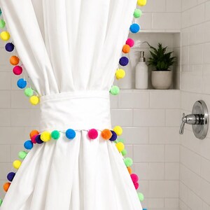 May include: White shower curtain with a colorful pom-pom trim. The curtain is hanging in a bathroom with white tile walls and a shower head. A small shelf holds a plant and soap dispenser.