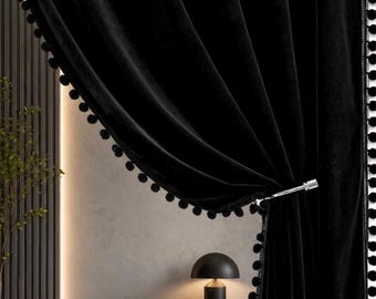 Può includere: Tende di velluto nero con finiture a pompon e fermatenda argentato. Le tende sono drappeggiate, rivelando un interno moderno con una consolle nera e una lampada nera.