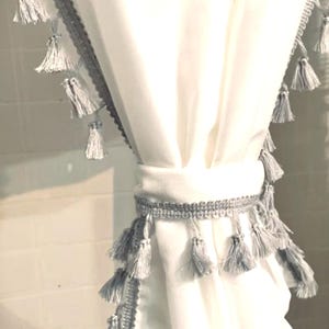 May include: White curtain with gray tassel trim and a matching tie-back. The curtain has a soft, flowing texture, and the tassels add a decorative element. The design is simple and elegant, suitable for home decor.