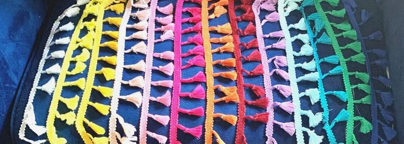 Pu&ograve; includere: Una collezione di strisce di passamaneria in tessuto disposte verticalmente con piccole nappe pendenti in uno spettro di colori arcobaleno che include bianco, giallo, pesca, rosa, magenta, arancione, rosso, viola chiaro, azzurro, verde e blu scuro.