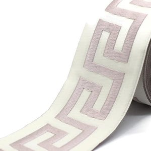 May include: A roll of white fabric ribbon featuring a repeating mauve Greek key pattern embroidered along the center. The ribbon has a smooth, satin-like texture and is wound around a circular cardboard core, showing the continuous geometric design.