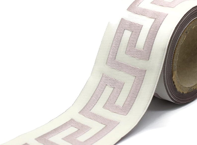May include: A roll of white fabric ribbon featuring a repeating mauve Greek key pattern embroidered along the center. The ribbon has a smooth, satin-like texture and is wound around a circular cardboard core, showing the continuous geometric design.
