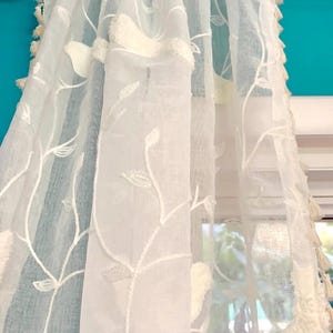 Pu&ograve; includere: Un pannello per tenda bianco semitrasparente con un motivo ricamato di viticci e foglie in filo bianco. Il tessuto ha una finitura testurizzata ed &egrave; decorato con piccole nappe bianche pendenti lungo i bordi verticali, appeso a un bastone.