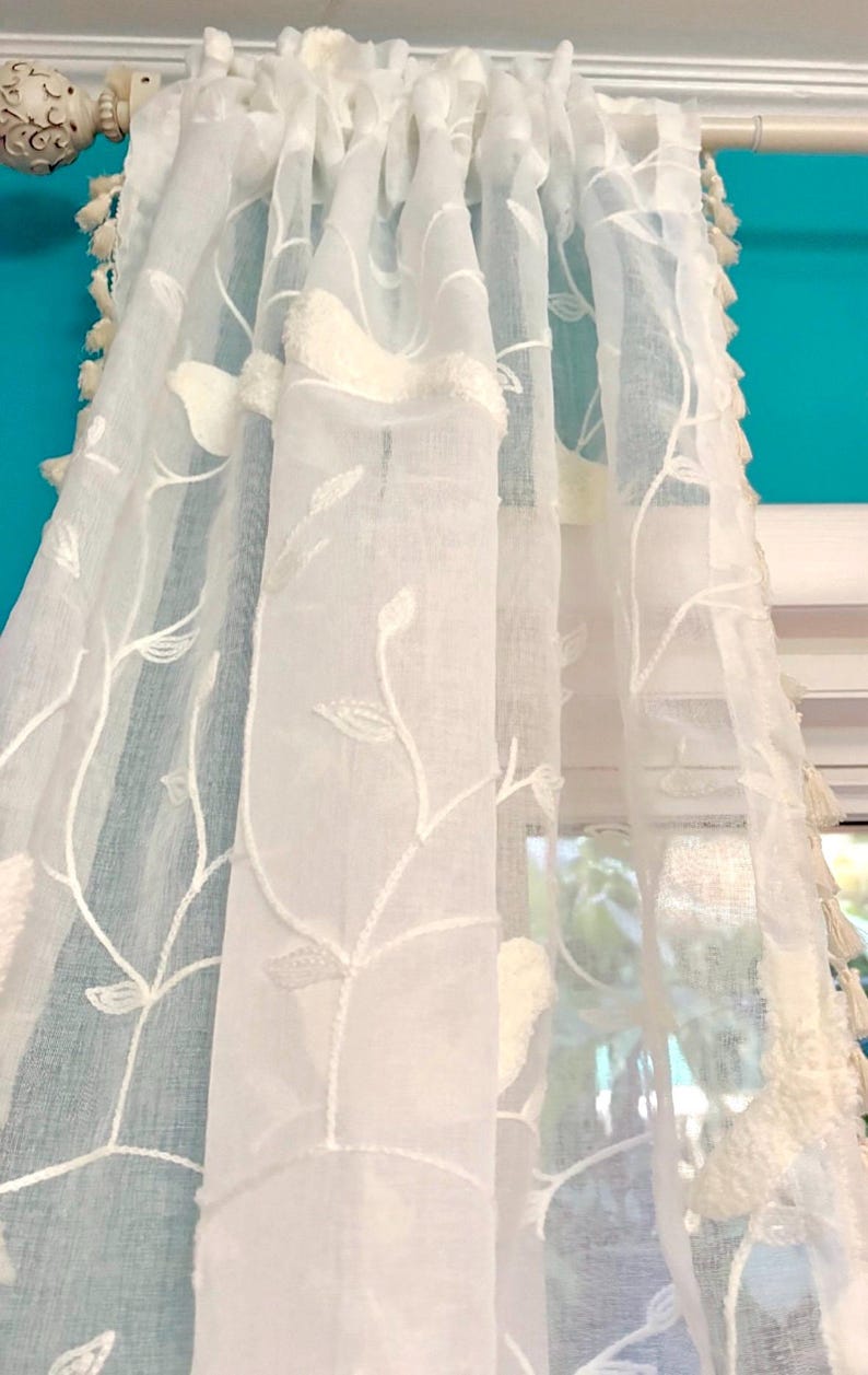 Pu&ograve; includere: Un pannello per tenda bianco semitrasparente con un motivo ricamato di viticci e foglie in filo bianco. Il tessuto ha una finitura testurizzata ed &egrave; decorato con piccole nappe bianche pendenti lungo i bordi verticali, appeso a un bastone.