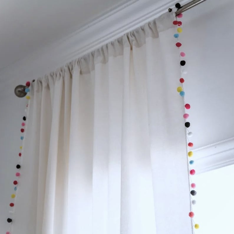 May include: White sheer curtains with a colorful pom-pom trim. The pom-poms are in various colors, including pink, yellow, blue, and black.