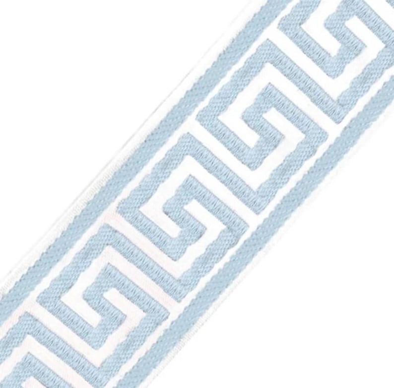 May include: A strip of fabric trim featuring a repeating light blue Greek key pattern embroidered onto a solid white background. The design consists of continuous interlocking geometric lines that run diagonally across the length of the trim.