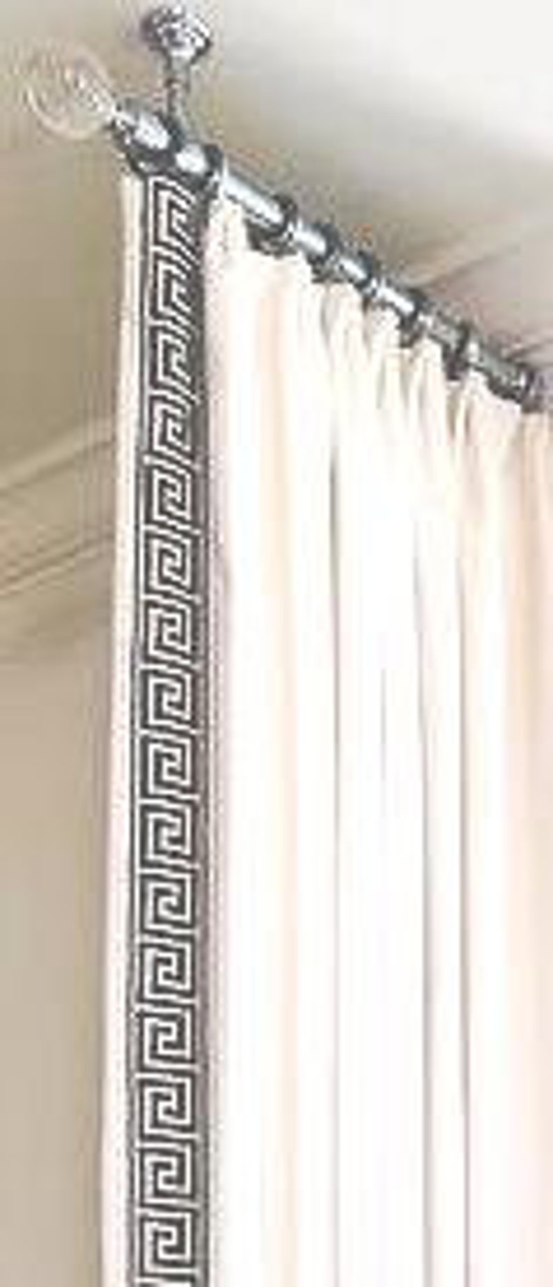May include: A cream-colored curtain hangs from a metallic rod with decorative finials. The curtain features a prominent vertical border along the leading edge, decorated with a repeating black and white Greek key geometric pattern.