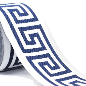 May include: A roll of woven fabric trim featuring a repeating dark blue Greek key pattern on a crisp white background. The ribbon has a textured surface and is wound around a central cardboard core, displaying a continuous geometric design.