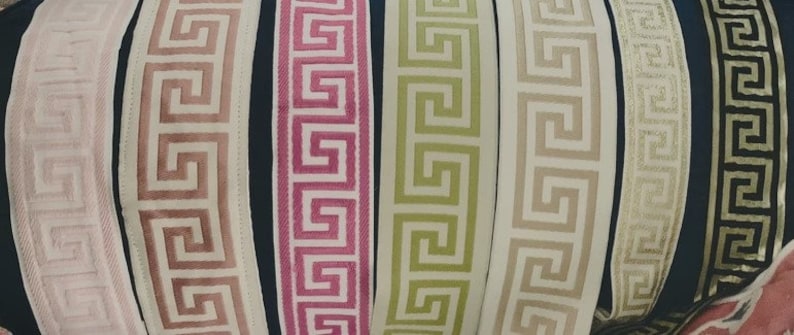May include: A collection of eight vertical fabric trim strips featuring a repeating Greek key pattern. The colors include pale pink, dusty rose, magenta, lime green, cream, beige, metallic gold, and black with gold, all arranged side by side.