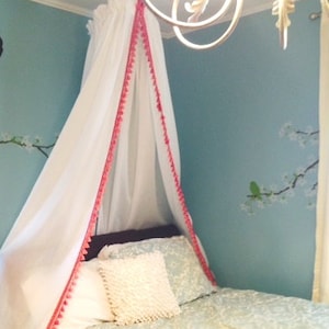 May include: A white fabric bed canopy featuring a bright pink pom-pom trim along the front edges. The canopy is suspended from the ceiling, draping down to cover the head of the bed, which is dressed in patterned light blue and white bedding.