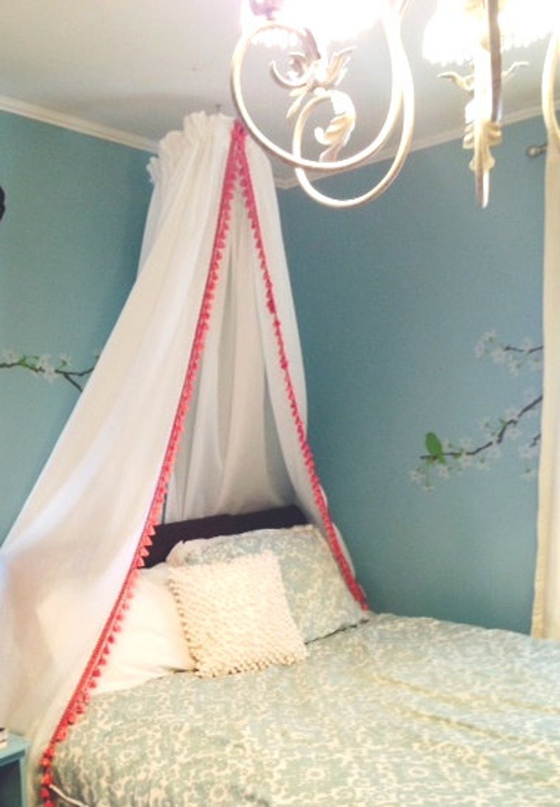 May include: A white fabric bed canopy featuring a bright pink pom-pom trim along the front edges. The canopy is suspended from the ceiling, draping down to cover the head of the bed, which is dressed in patterned light blue and white bedding.