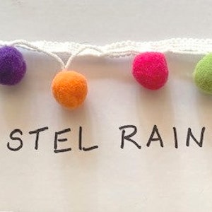 May include: A white woven trim featuring a series of small, fuzzy pom-poms in various colors including lime green, bright blue, purple, orange, and hot pink. The text "PASTEL RAINBOW" is written in black ink below the colorful pom-pom trim.