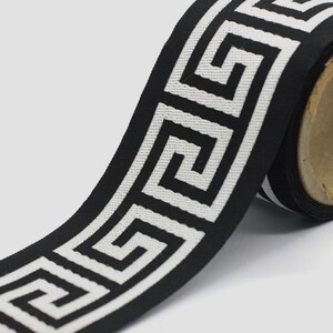 May include: A roll of black fabric ribbon featuring a repeating white Greek key geometric pattern. The woven trim is wound around a cylindrical cardboard core, displaying a continuous linear design with sharp, angular, interlocking lines.