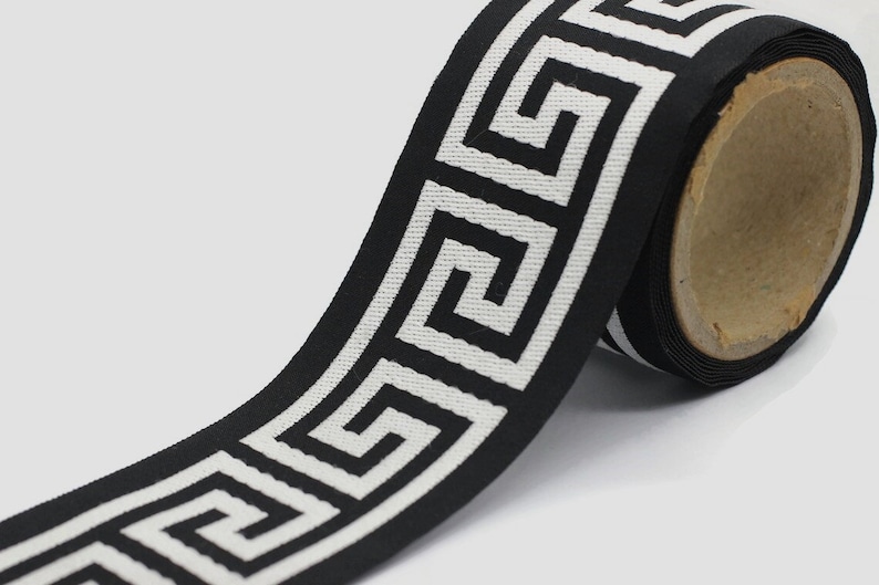 May include: A roll of black fabric ribbon featuring a repeating white Greek key geometric pattern. The woven trim is wound around a cylindrical cardboard core, displaying a continuous linear design with sharp, angular, interlocking lines.
