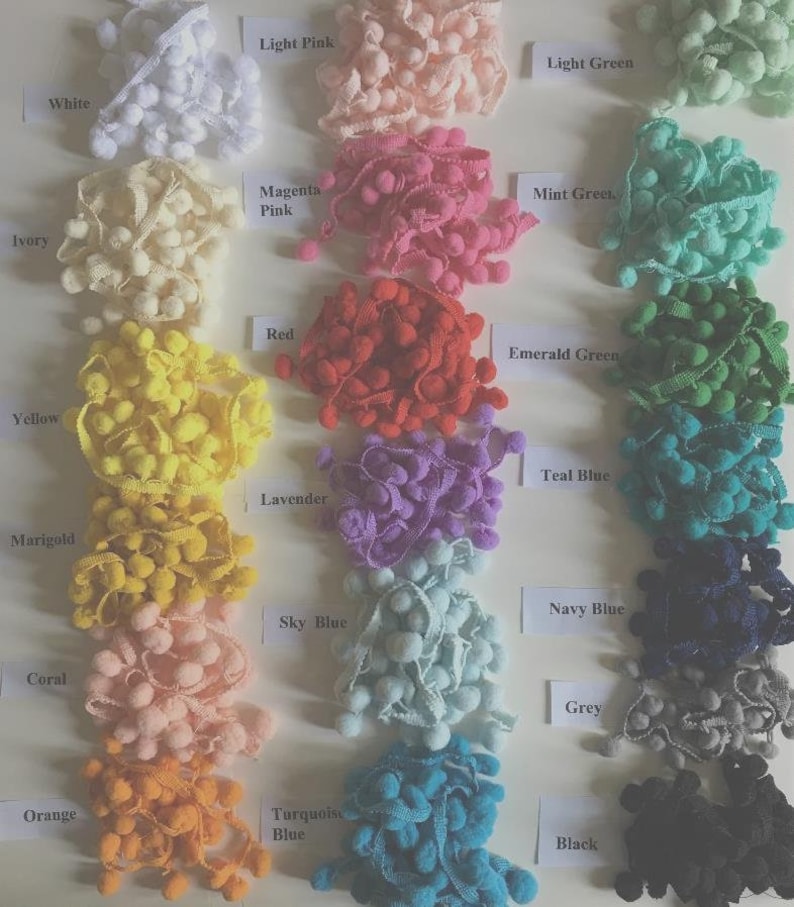 May include: A collection of pom-pom trim organized in three vertical columns, featuring various colors labeled with white paper tags including white, ivory, yellow, marigold, coral, orange, light pink, magenta pink, red, lavender, sky blue, turquoise blue, light green, mint green, emerald green, teal blue, navy blue, grey, and black.