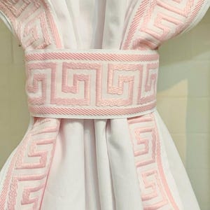 May include: A white curtain panel gathered in the center by a matching tieback. The curtain and tieback feature a wide, light pink embroidered trim with a repeating geometric Greek key pattern along the edges and the center band.