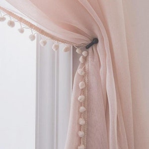 May include: A sheer pink curtain with a white pom-pom trim. The curtain is tied back with a black clip.