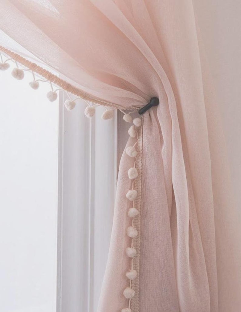 May include: A sheer pink curtain with a white pom-pom trim. The curtain is tied back with a black clip.