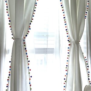 May include: White curtains with a colorful pom-pom trim. The curtains are drawn back to reveal a window with white blinds.