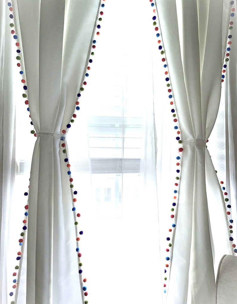May include: White curtains with a colorful pom-pom trim. The curtains are drawn back to reveal a window with white blinds.