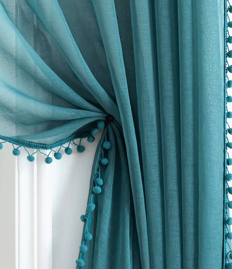 May include: Teal sheer curtains with a pom-pom trim. The curtains are gathered at the top and tied back with a matching ribbon.
