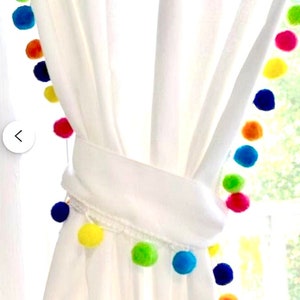 May include: White curtain tied back with a matching fabric tie, decorated with a colourful pom-pom garland. The pom-poms are in shades of red, orange, yellow, green, and blue, adding a playful touch to the window treatment.