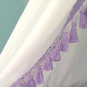 May include: A white fabric curtain featuring a decorative trim edge adorned with a series of light purple tassels. The trim consists of a woven band from which individual, uniform lavender-colored thread tassels hang at regular intervals.