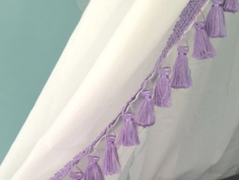 May include: A white fabric curtain featuring a decorative trim edge adorned with a series of light purple tassels. The trim consists of a woven band from which individual, uniform lavender-colored thread tassels hang at regular intervals.