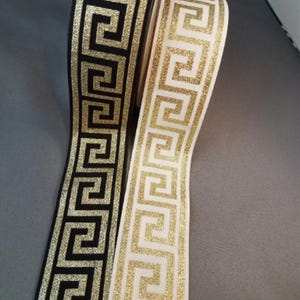 May include: Two rolls of ribbon featuring a repeating gold glitter Greek key pattern. One roll has a black background, and the other roll has a white background. The ribbons are displayed vertically, showing the continuous geometric design.