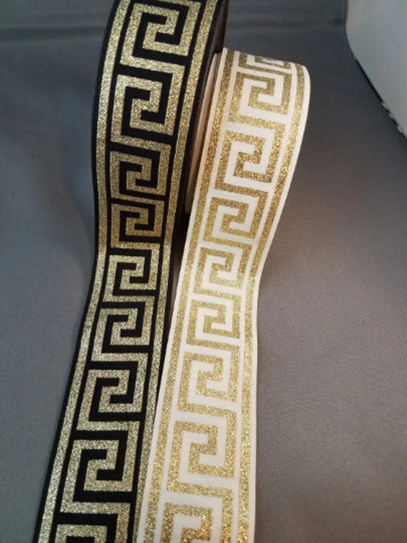 May include: Two rolls of ribbon featuring a repeating gold glitter Greek key pattern. One roll has a black background, and the other roll has a white background. The ribbons are displayed vertically, showing the continuous geometric design.