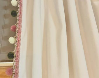 Può includere: Una tenda color crema con pieghe verticali e un bordo decorativo lungo il lato. Il bordo è costituito da una passamaneria intrecciata rosa e verde decorata con una serie di pompon alternati in verde scuro, rosa chiaro e bianco.