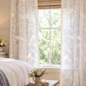 Pu&ograve; includere: Una coppia di tende bianche semitrasparenti con un motivo ricamato di uccelli appollaiati su rami. Le tende sono decorate con piccole nappe bianche lungo i bordi verticali interni e sono appese a un bastone nero sopra una finestra.