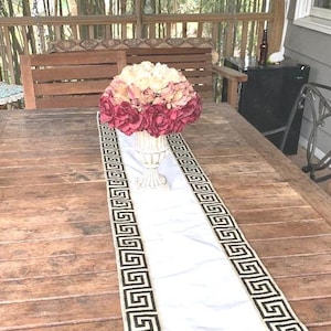 May include: A white table runner with a black Greek key pattern border. A white vase with a pink and cream flower arrangement sits on the runner.