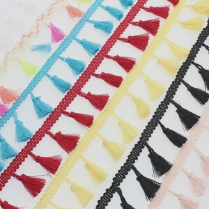May include: A collection of decorative tassel fringe trim strips arranged diagonally. The trim features small, hanging tassels in various solid colors including bright blue, deep red, pale yellow, black, and soft pink, attached to a woven header.