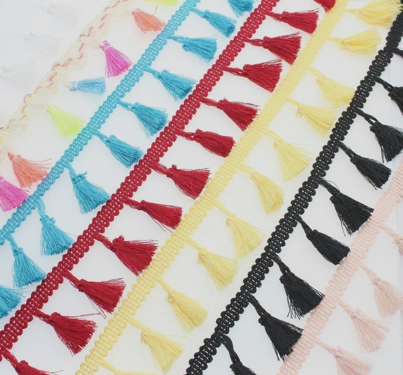 May include: A collection of decorative tassel fringe trim strips arranged diagonally. The trim features small, hanging tassels in various solid colors including bright blue, deep red, pale yellow, black, and soft pink, attached to a woven header.