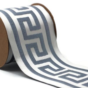 May include: A spool of white ribbon featuring a repeating gray Greek key pattern. The ribbon is wound around a circular brown cardboard core, with the patterned fabric extending outward to display the geometric design clearly.